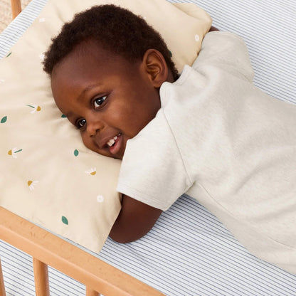 Smiling toddler resting face down on a beige pillow decorated with daisies and green leaves inside a wooden cot.