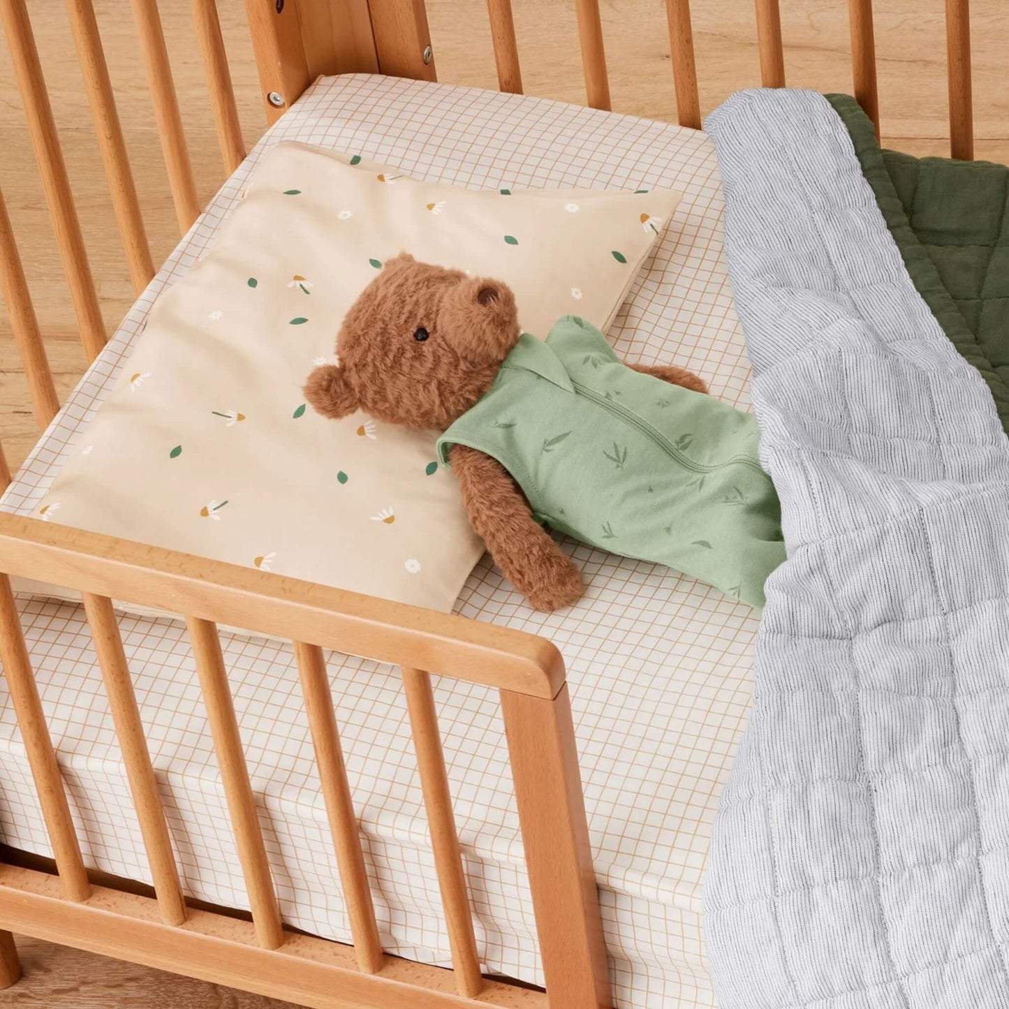Stuffed teddy bear tucked under a blanket in a cot, head resting on a beige pillow printed with daisy flowers and leaves.