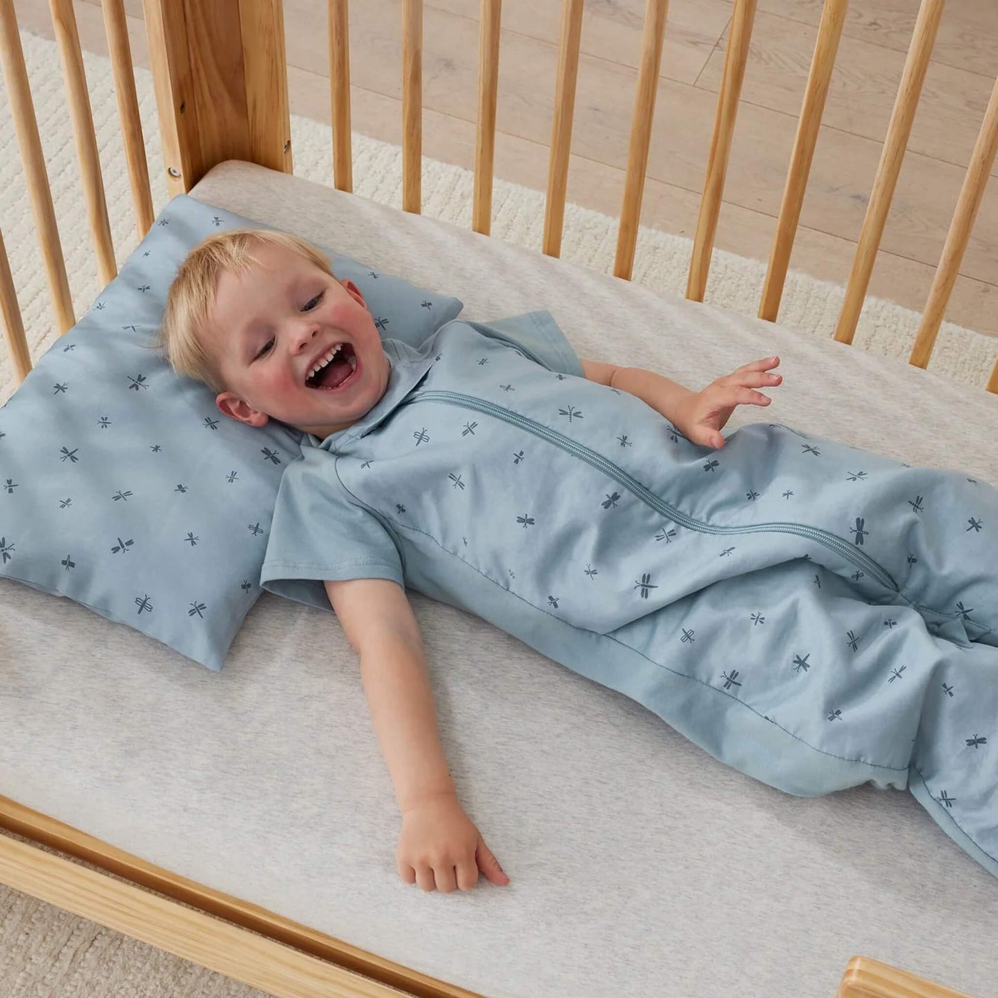 Smiling toddler lying on a light blue pillow patterned with dragonflies, wearing a matching sleep suit inside a wooden cot.