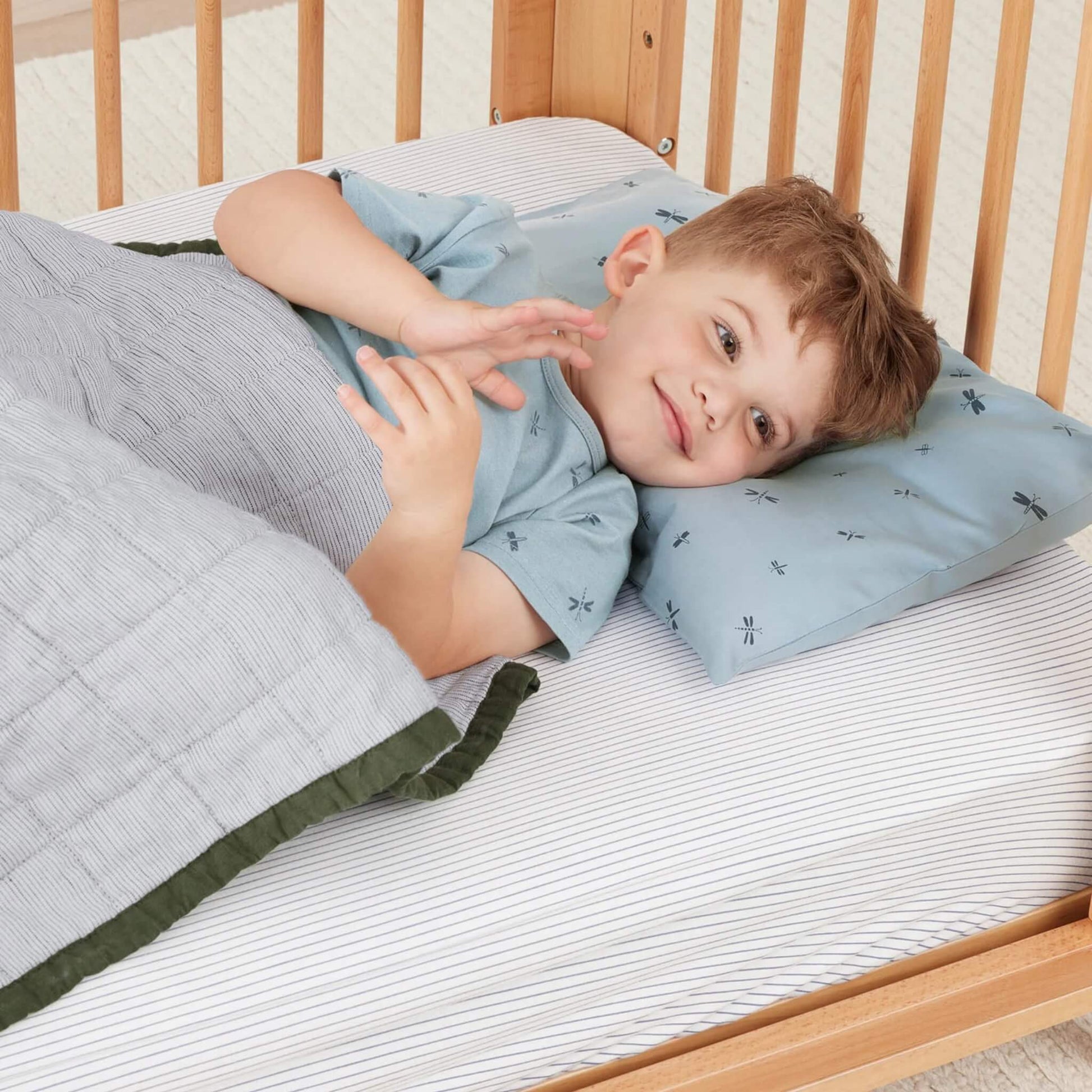 Toddler lying under a striped blanket in a cot, resting on a light blue pillow decorated with navy dragonfly illustrations.