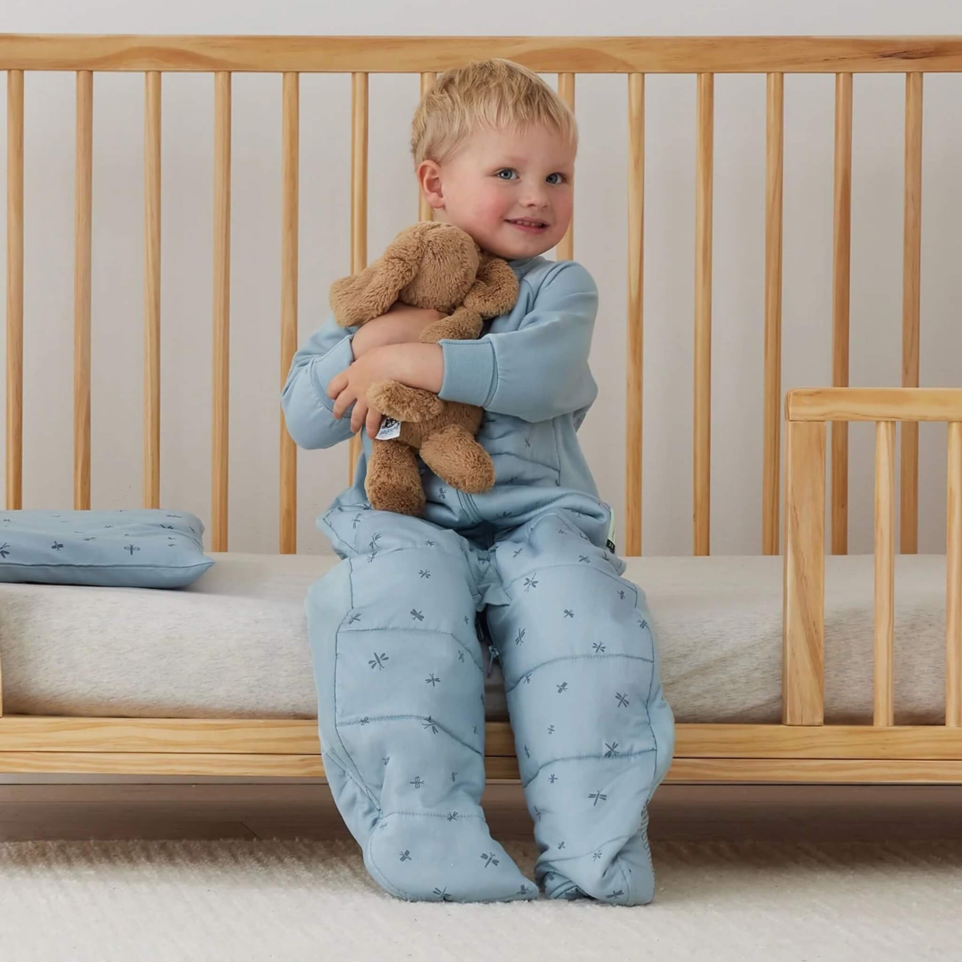 Toddler sitting on the edge of a cot, cuddling a soft toy with a folded light blue dragonfly pillow visible beside them.