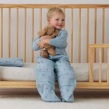 Toddler sitting on the edge of a cot, cuddling a soft toy with a folded light blue dragonfly pillow visible beside them.