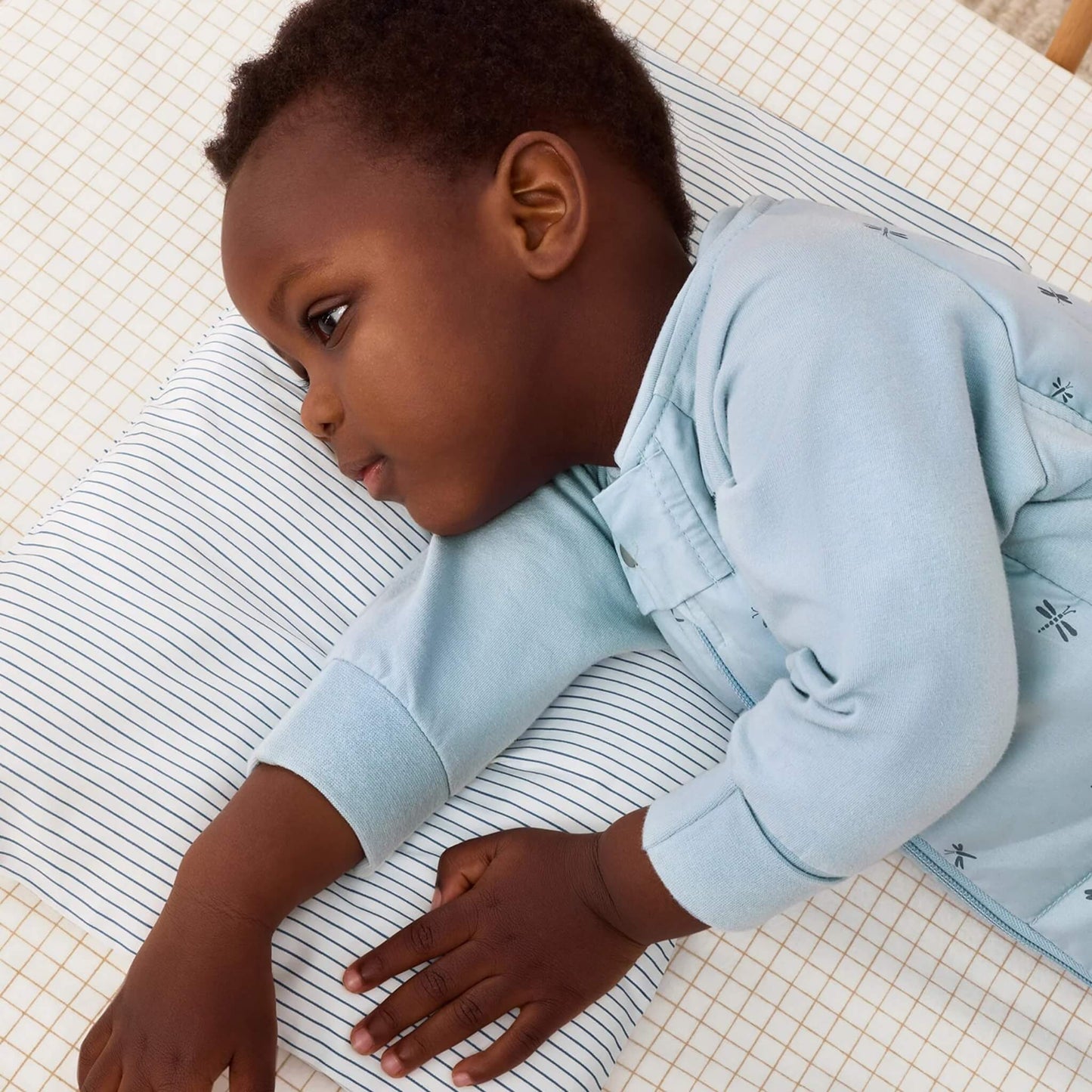 Toddler in a pale blue sleepsuit resting on a white pillow decorated with navy stripes inside a cot.