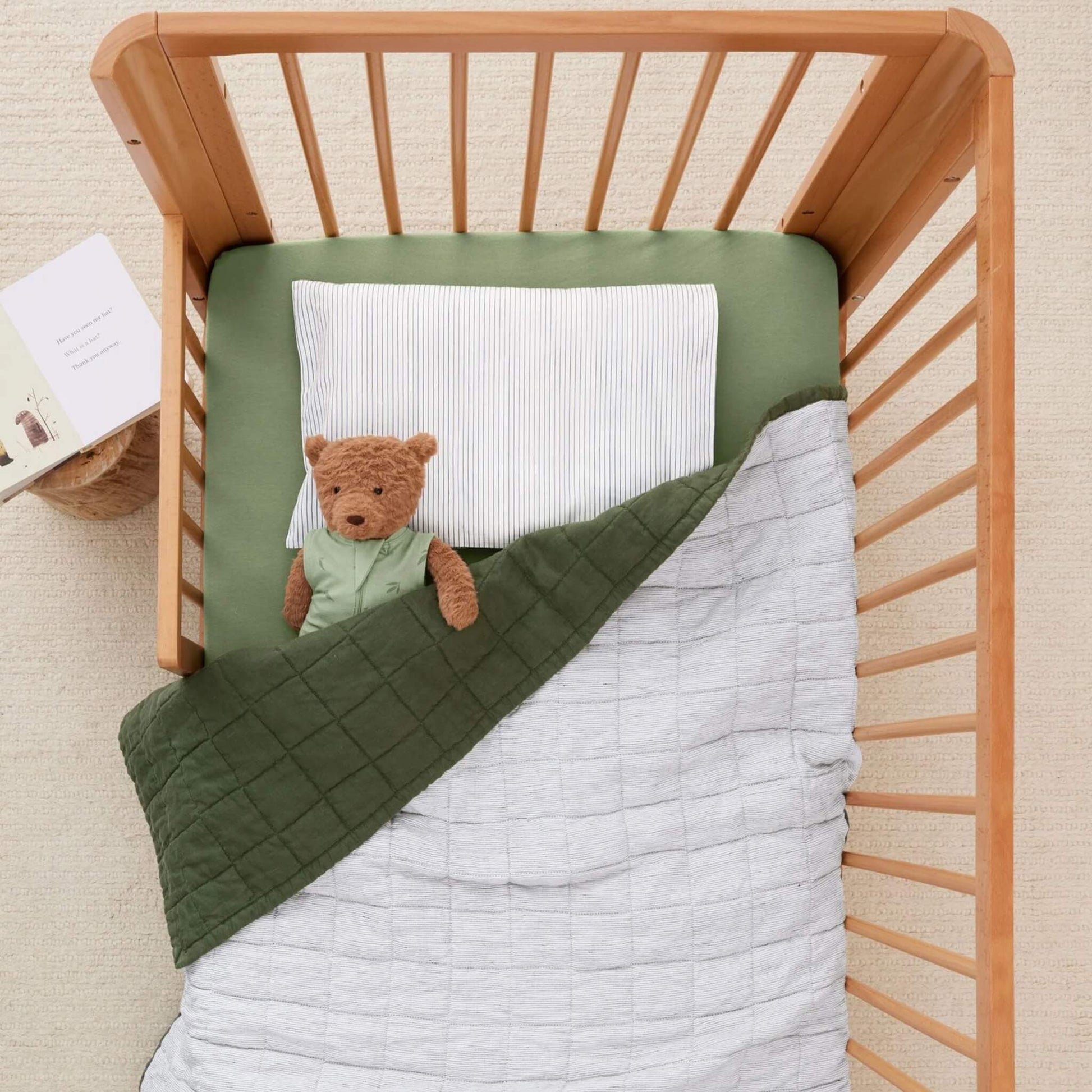 Stuffed teddy bear tucked under a quilt in a cot, head resting on a striped white and navy toddler pillow.