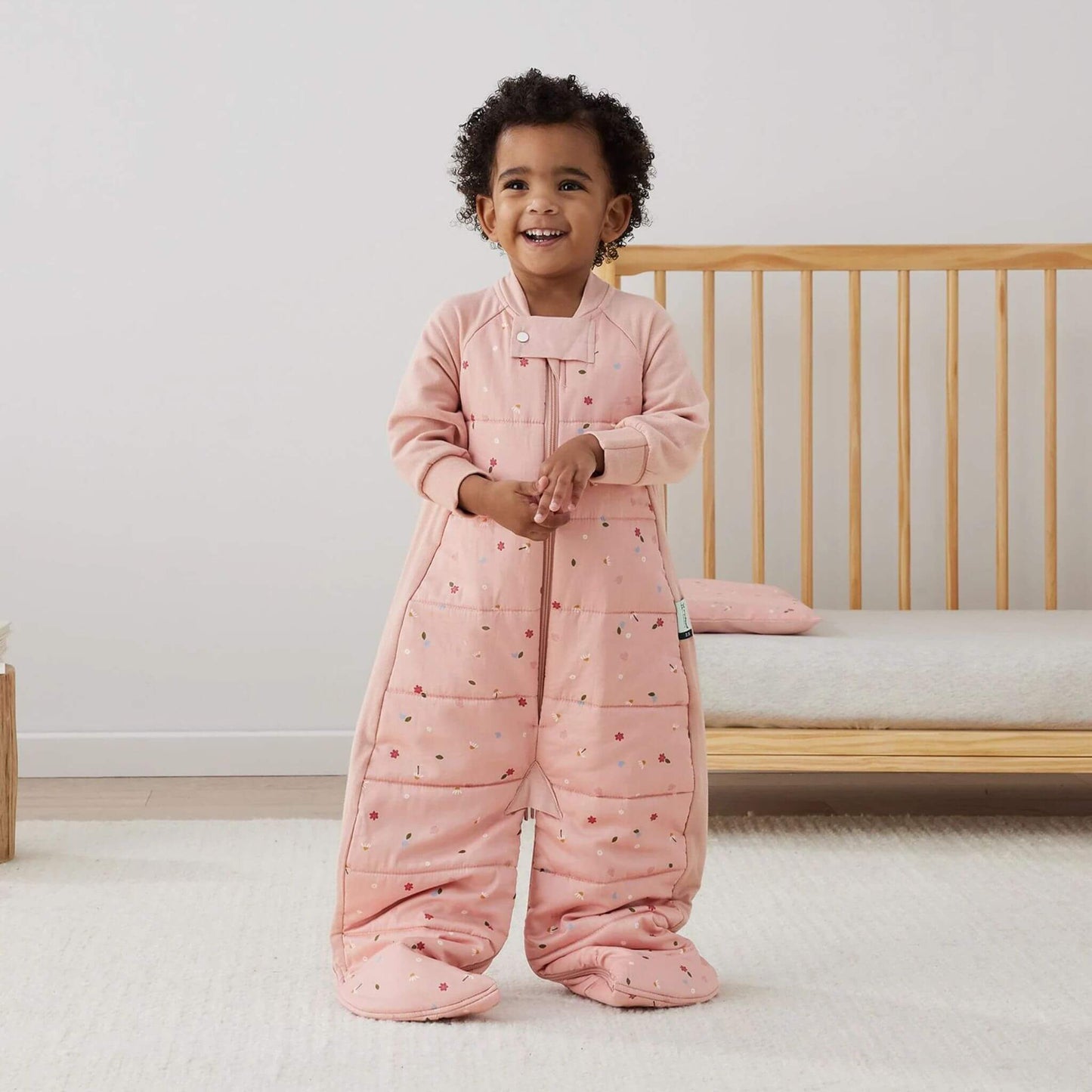 Toddler standing beside a cot wearing a sleep suit bag in pink daisies print, shown in sleep suit mode with feet covered.