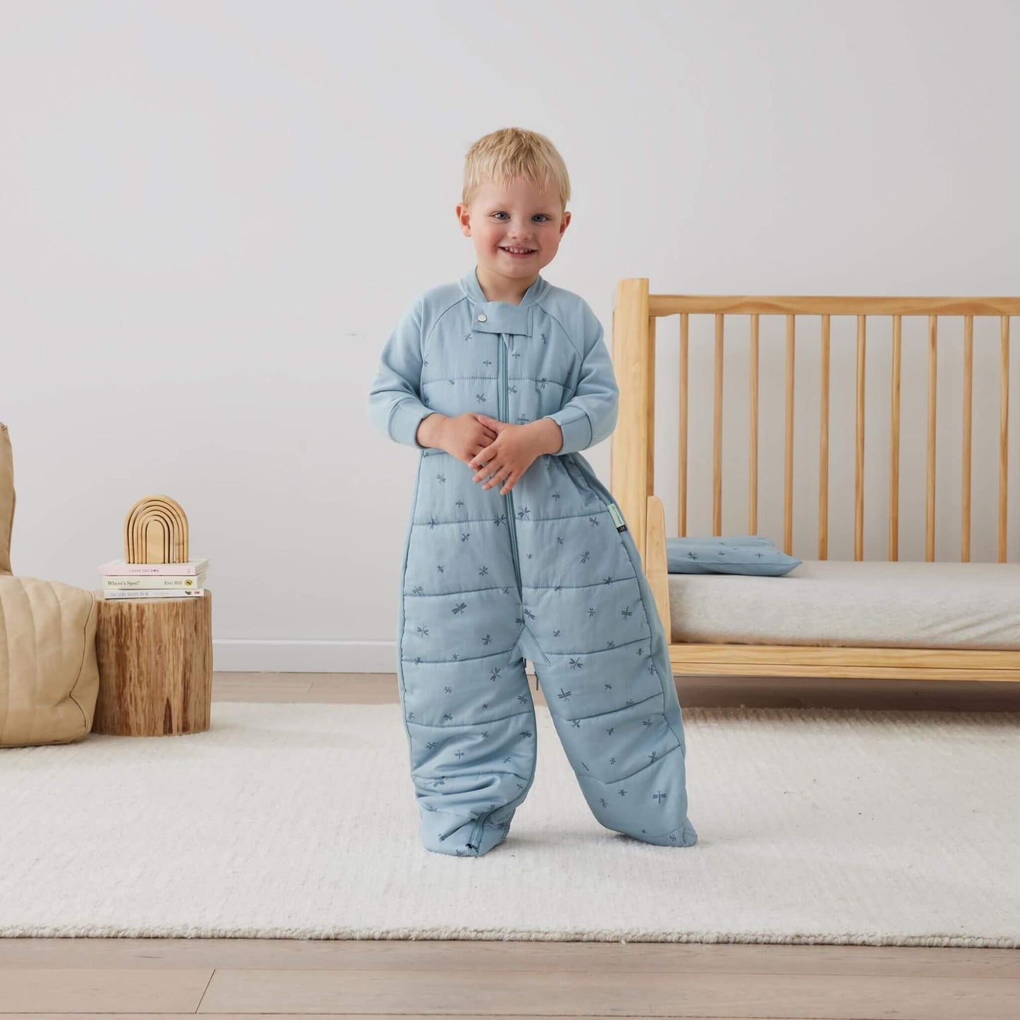 Smiling child standing beside a cot while wearing the ergoPouch sleep suit bag in blue dragonflies print with separated legs.