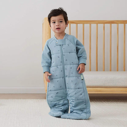 Young child standing in a nursery wearing a blue dragonflies sleep suit bag, zipped in sleeping bag style with feet enclosed.