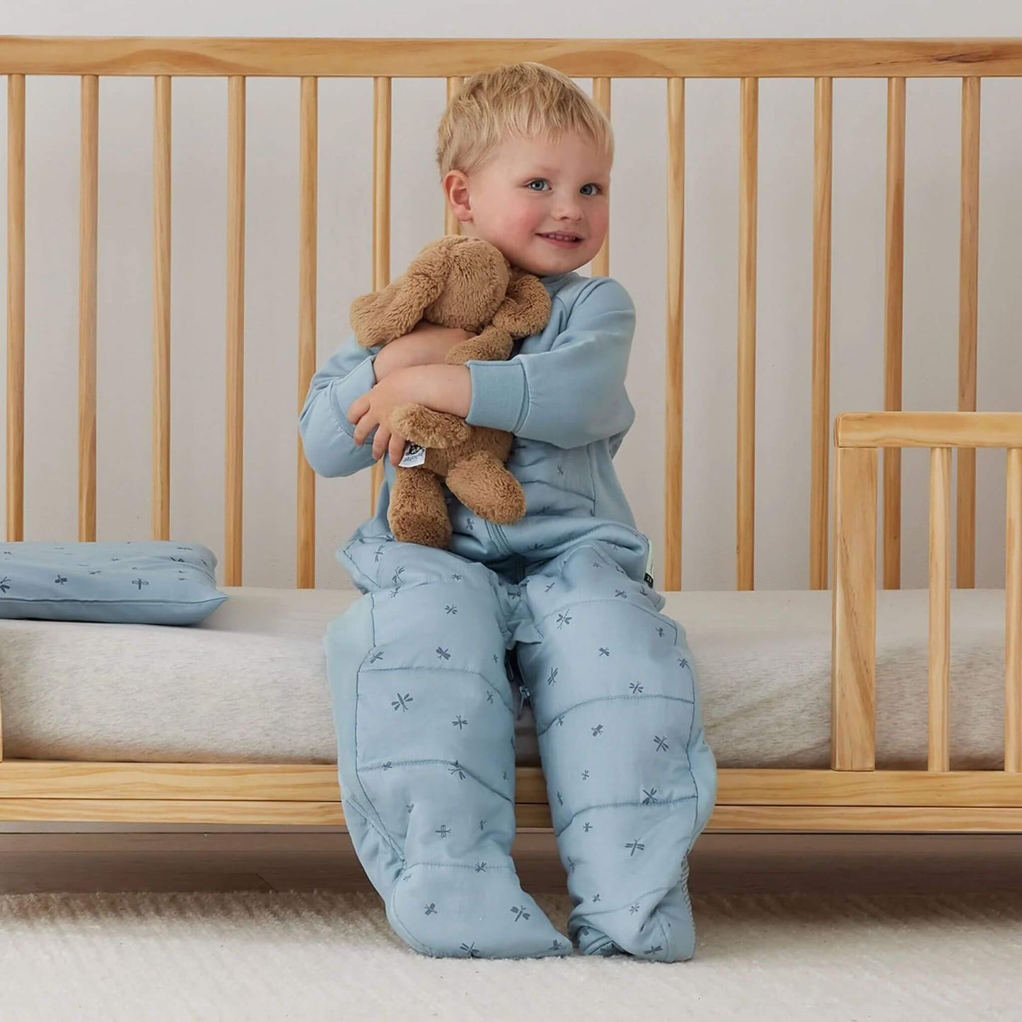 Child sitting inside a wooden cot hugging a teddy while dressed in a blue dragonflies print sleep suit bag with separated legs.