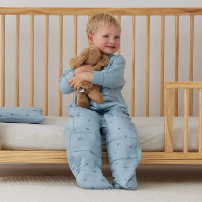 Child sitting inside a wooden cot hugging a teddy while dressed in a blue dragonflies print sleep suit bag with separated legs.