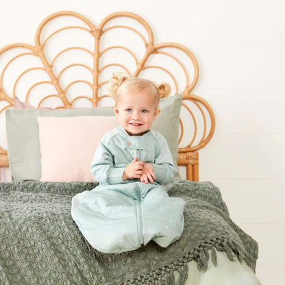 Toddler sitting on a bed wearing a padded sage sleep suit bag with zips closed.
