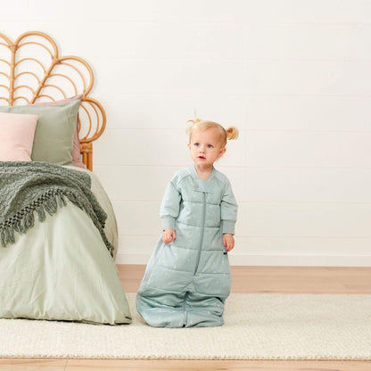 Toddler standing beside a bed wearing a sage padded sleep suit bag in sleeping bag shape.