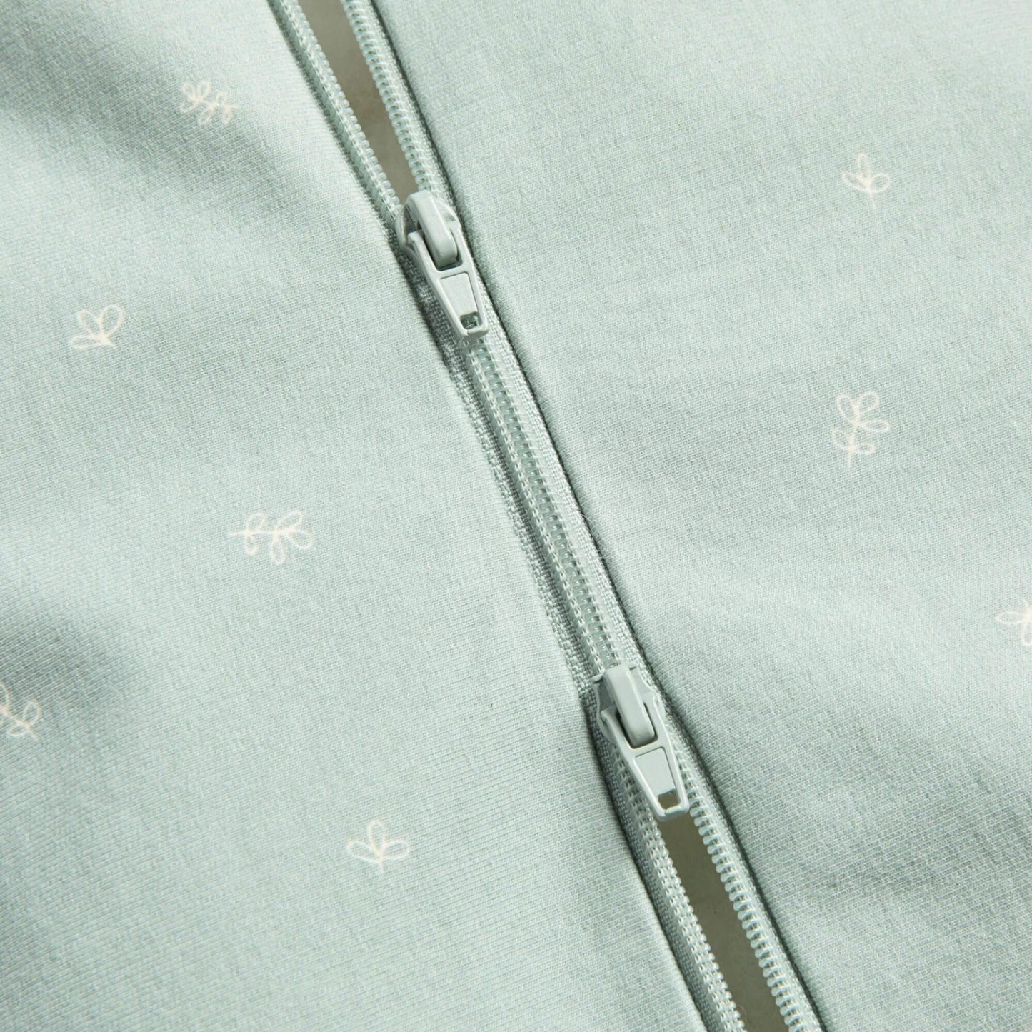 Close-up of a sage green baby Swaddle Bag showing a two-way zip detail with small white motifs on the fabric.