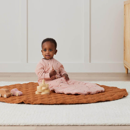 Toddler sitting comfortably wearing a pink long sleeved sleeping bag with Daisies print, playing on a soft quilted mat.