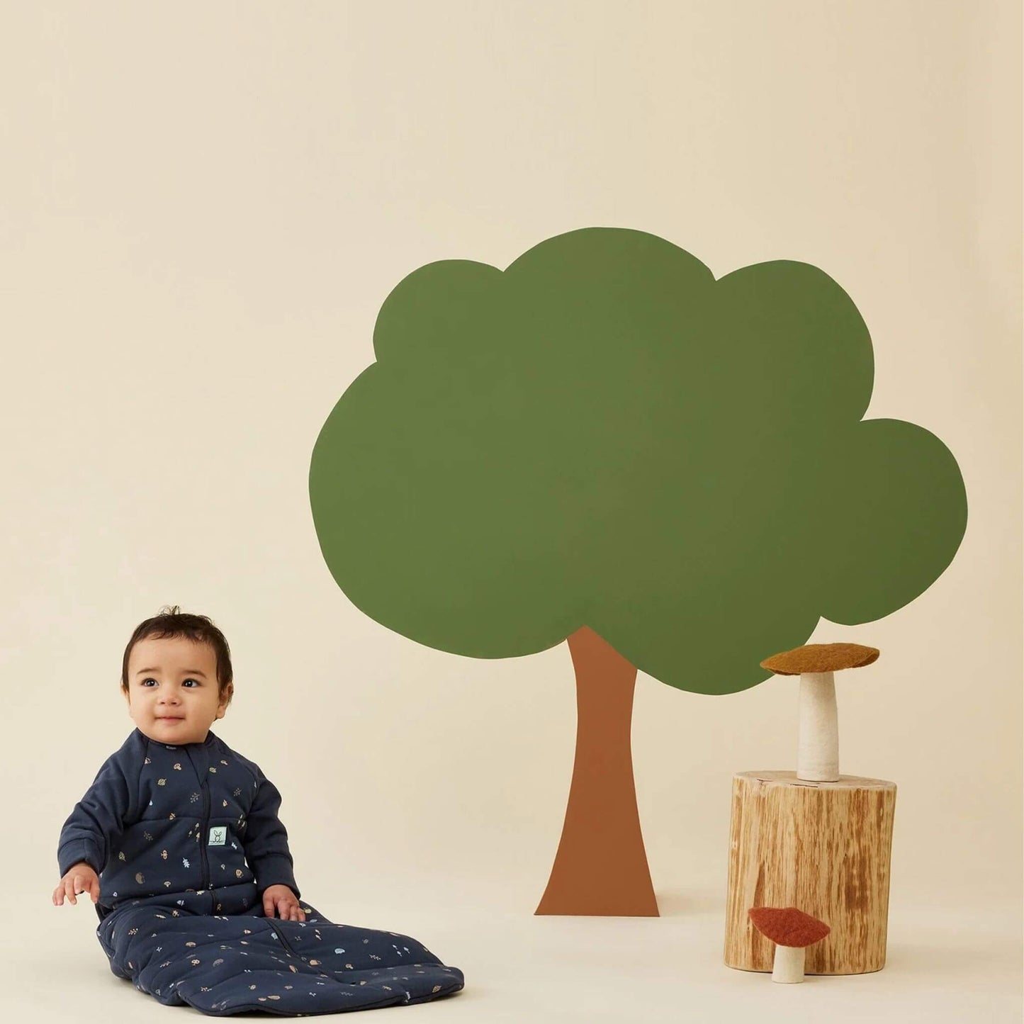 Toddler sitting in a long sleeved, winter weight sleeping bag in Hedgehog print, with a woodland-themed background featuring a tree and toadstools.