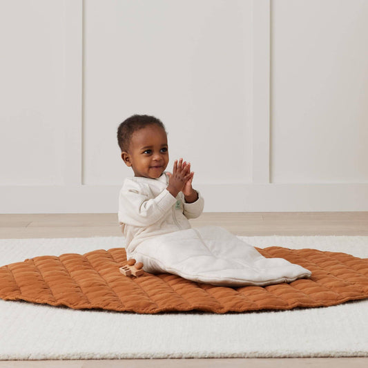 Toddler sitting happily in a Long Sleeved Sleeping Bag in Oatmeal, playing on a cosy quilted mat with wooden toys.