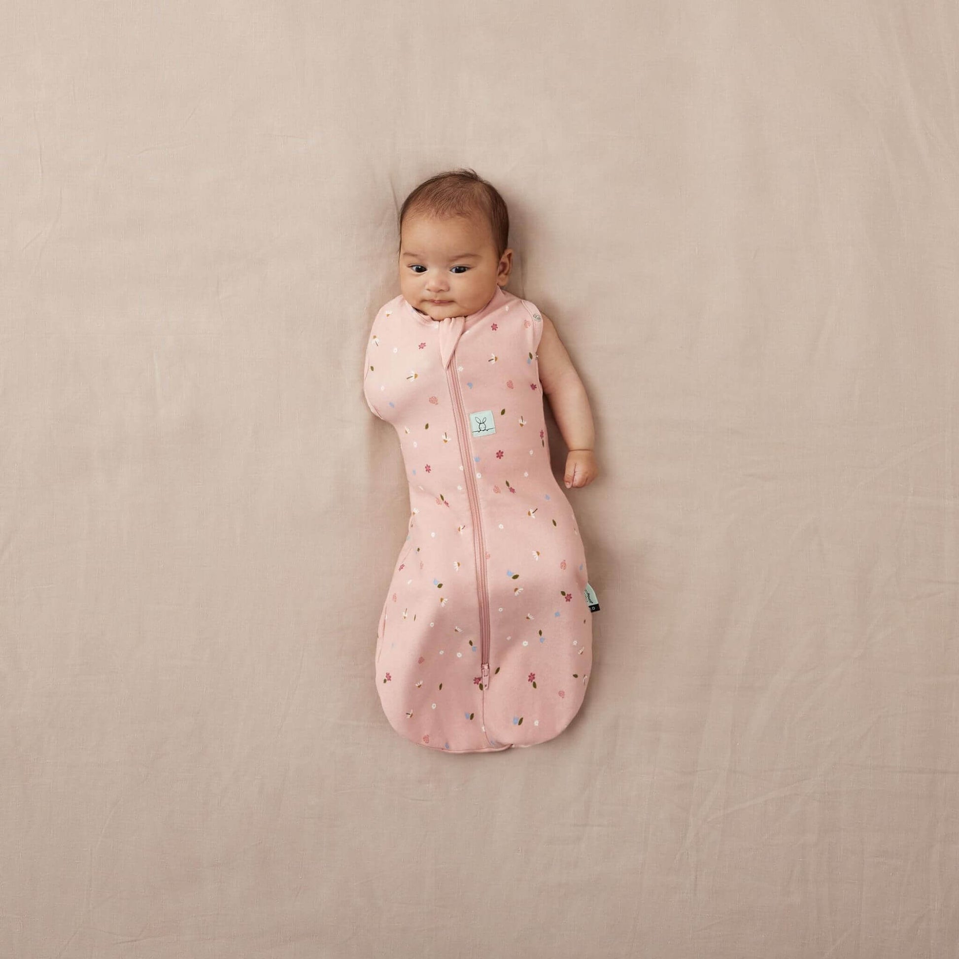 A baby lying snugly, with one arm out, in a swaddle Bag with a daisies print, designed for comfort, warmth, and safe sleep.