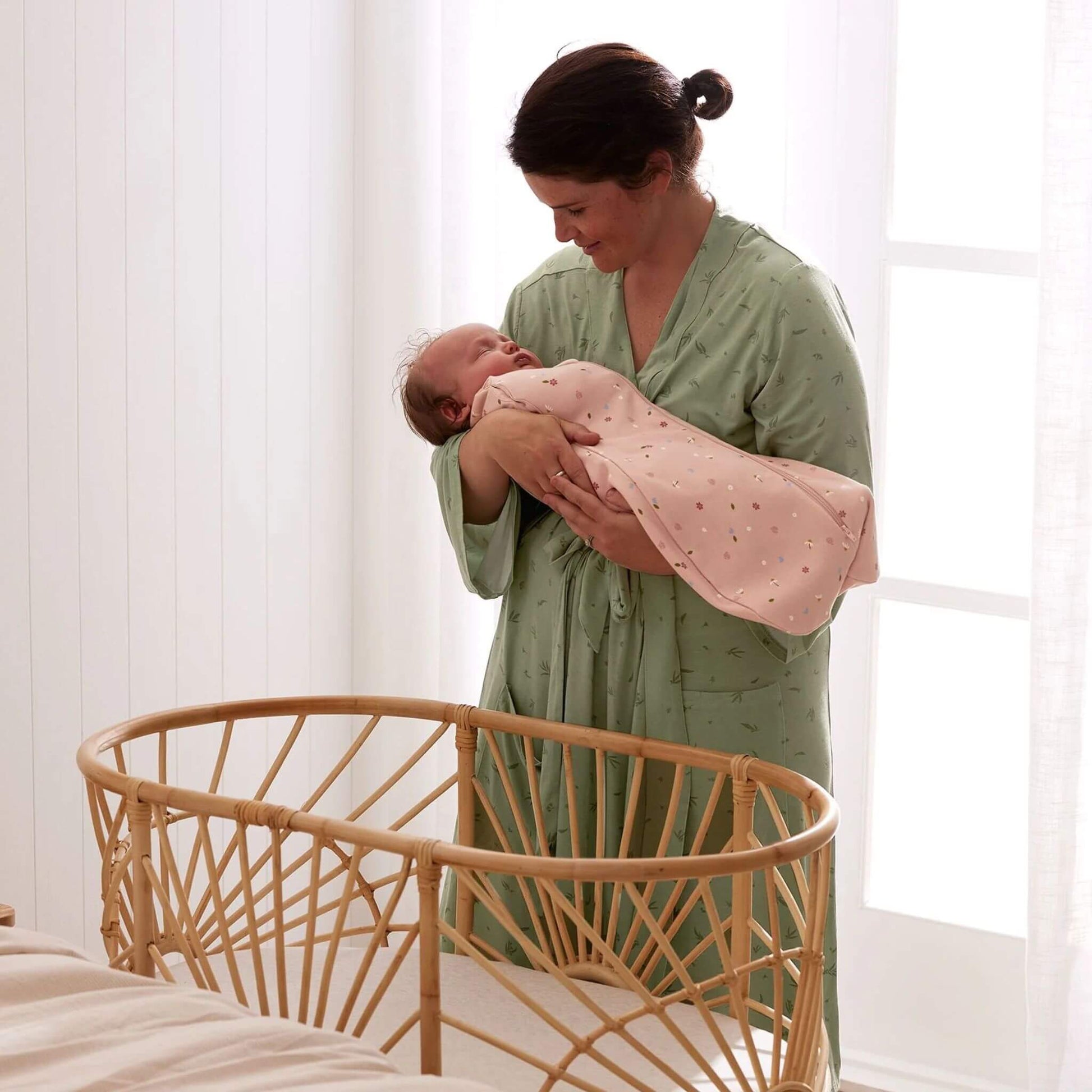A mother gently cradling her sleeping baby wrapped in a Swaddle Bag with a daisies print, near a stylish bassinet.