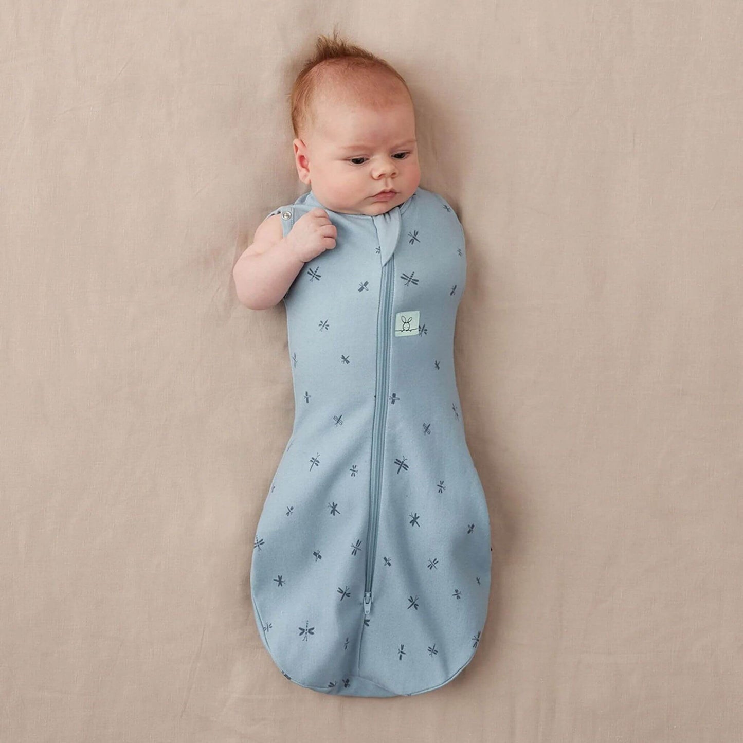 A baby wearing a Swaddle Bag in dragonflies print, transitioning with arm-out option for added comfort and mobility.