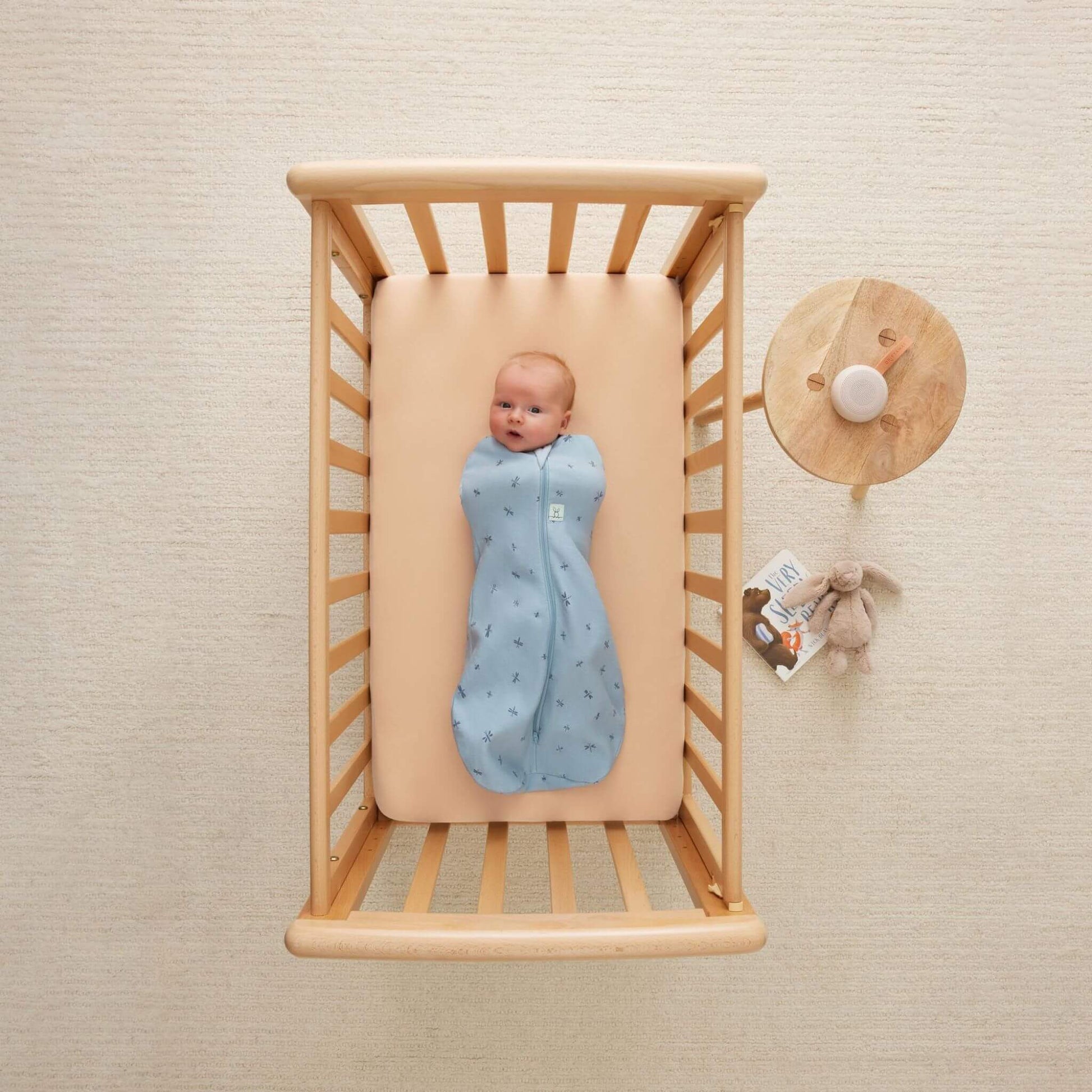 A baby lying snugly in a wooden cot, wearing a Swaddle Bag in dragonflies print, surrounded by soft nursery accents.