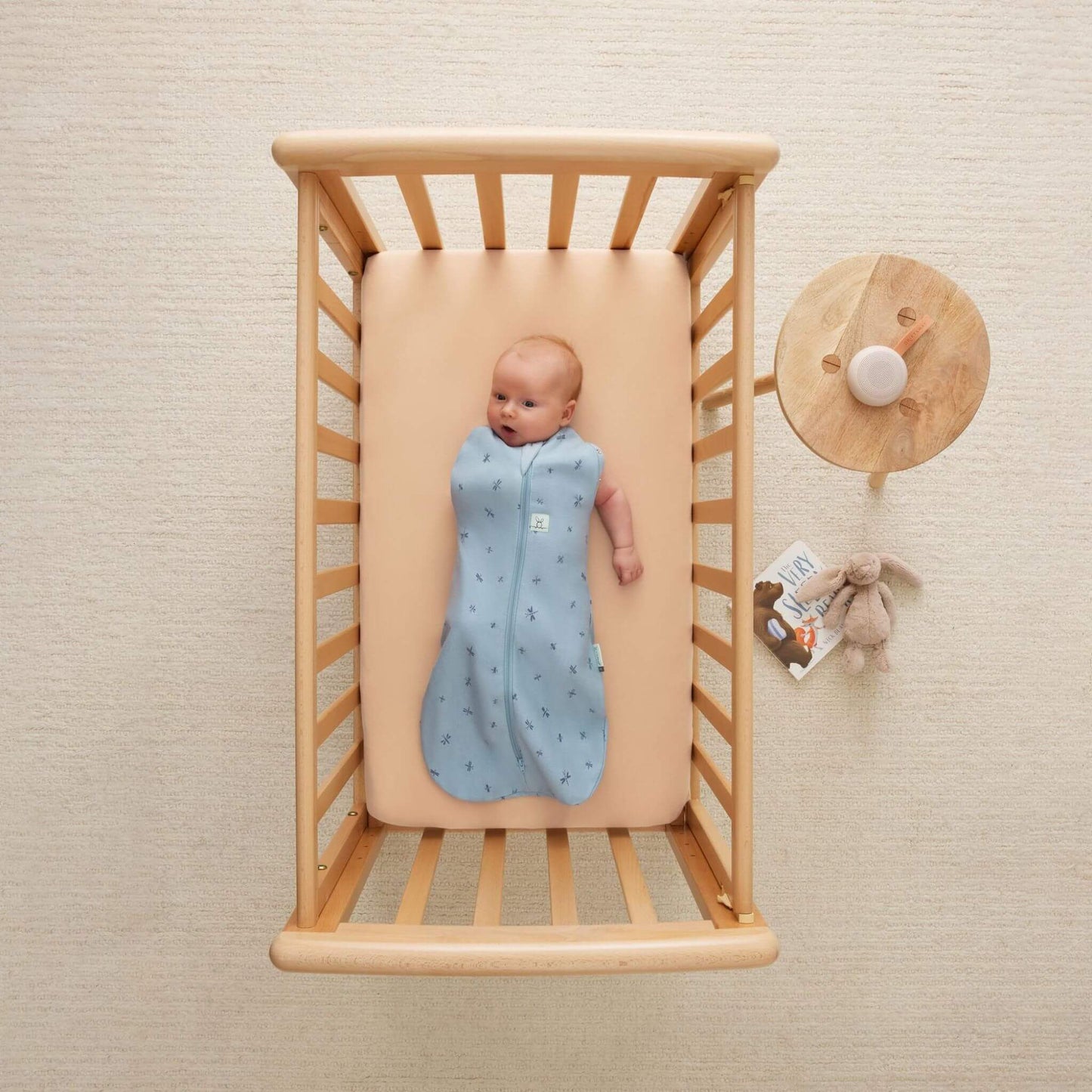 Baby lying in a wooden cot wearing a light blue Cocoon Swaddle Bag with dragonfly print, both arms free, with a soft toy and book placed nearby.