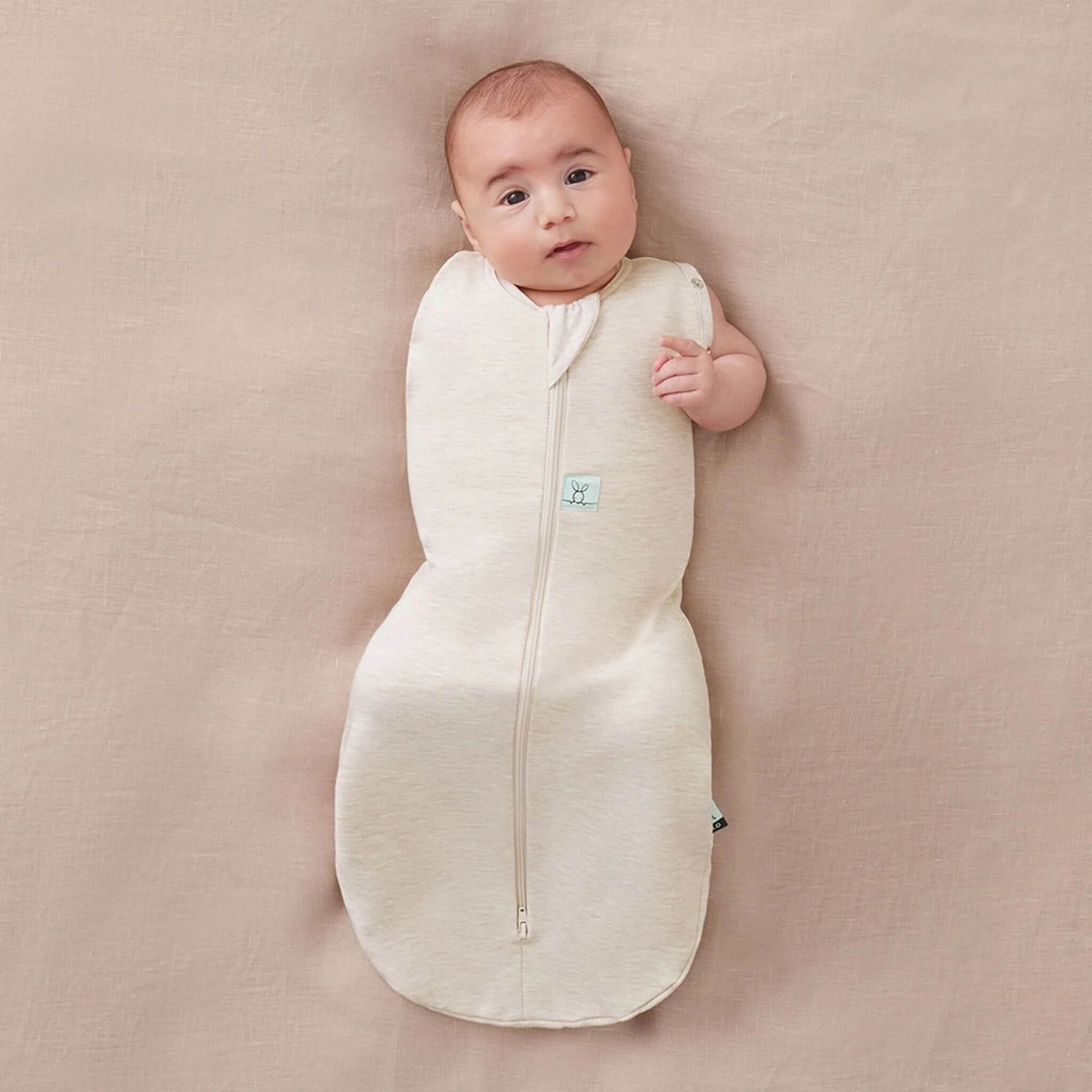 Baby comfortably resting in Cocoon Swaddle Bag in oatmeal, showcasing its arm-out option for flexible and safe sleeping.