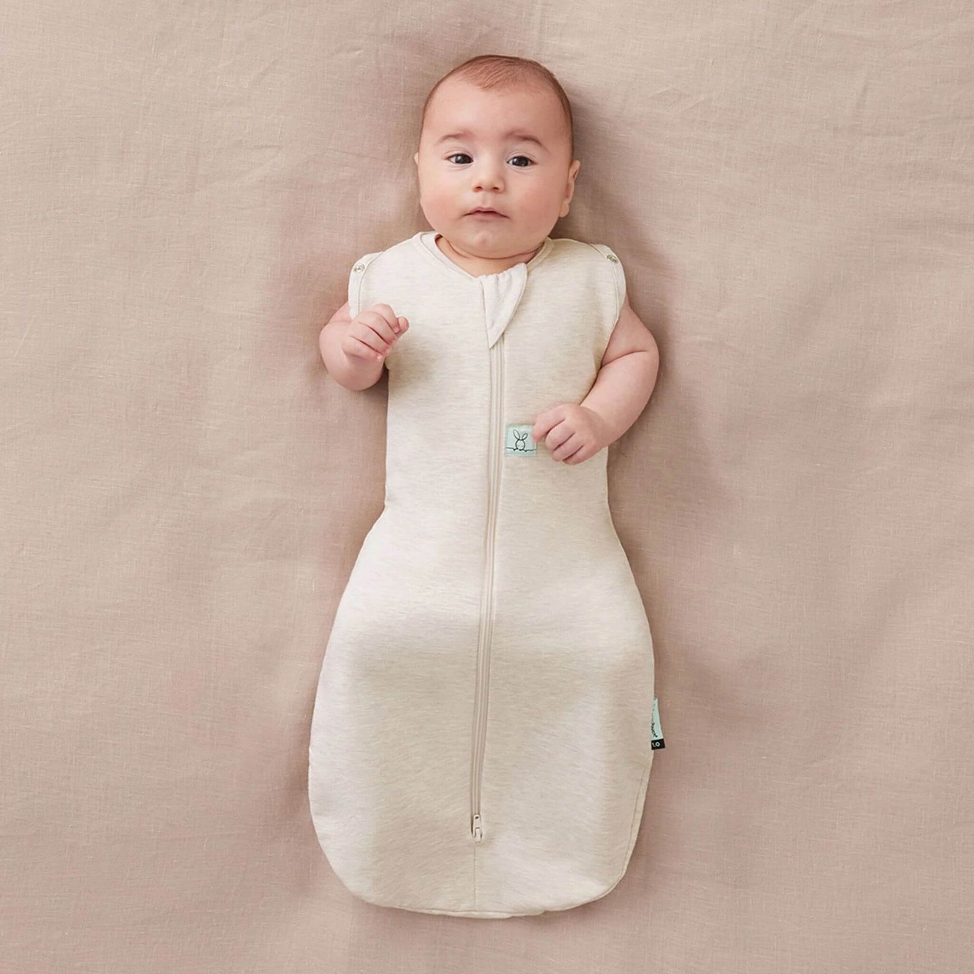 Baby lying comfortably in a Swaddle Bag in oatmeal, showcasing its breathable fabric and flexible arm-out sleeping option.
