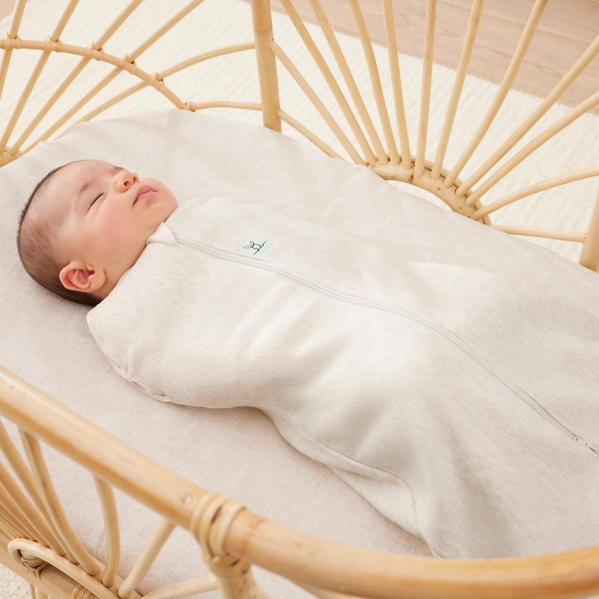 Baby peacefully sleeping in a rattan bassinet, wrapped in a Cocoon Swaddle Bag in oatmeal, offering snug comfort and security.