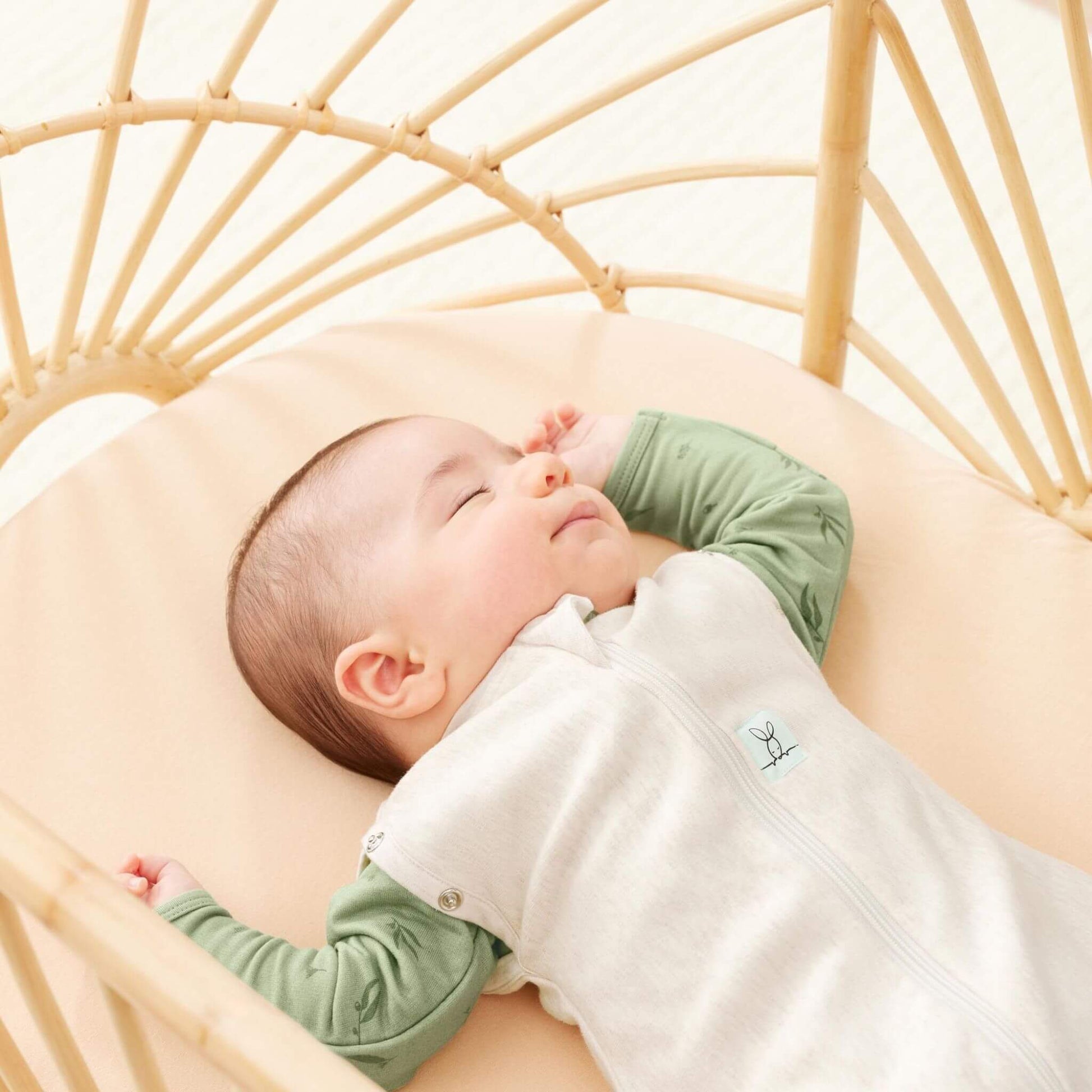 Baby sleeping peacefully in a rattan bassinet, wearing a Cocoon Swaddle Bag in oatmeal paired with a green underlayer.