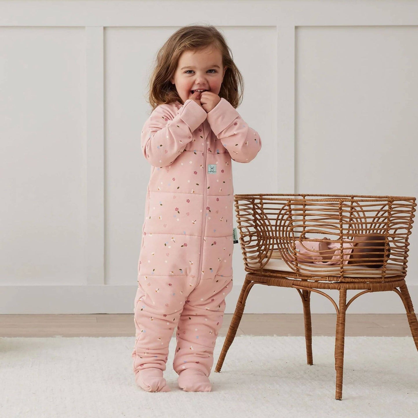 A toddler stands wearing a winter weight Sleep Onesie in the Daisies print. The Onesie, with its soft pink fabric with dainty floral pattern, offers cosy warmth and flexibility.