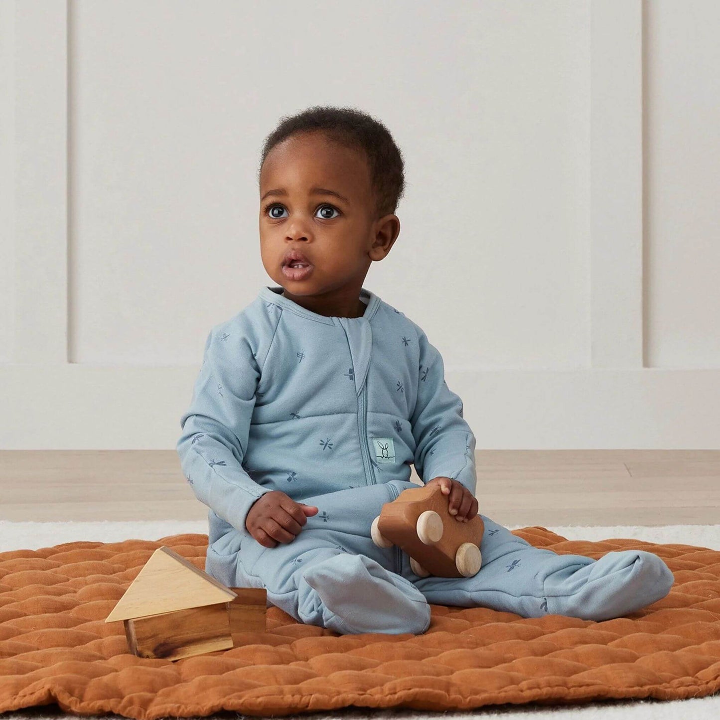 A toddler sitting comfortably on a quilted mat, wearing a blue Sleep Onesie in Dragonflies print.