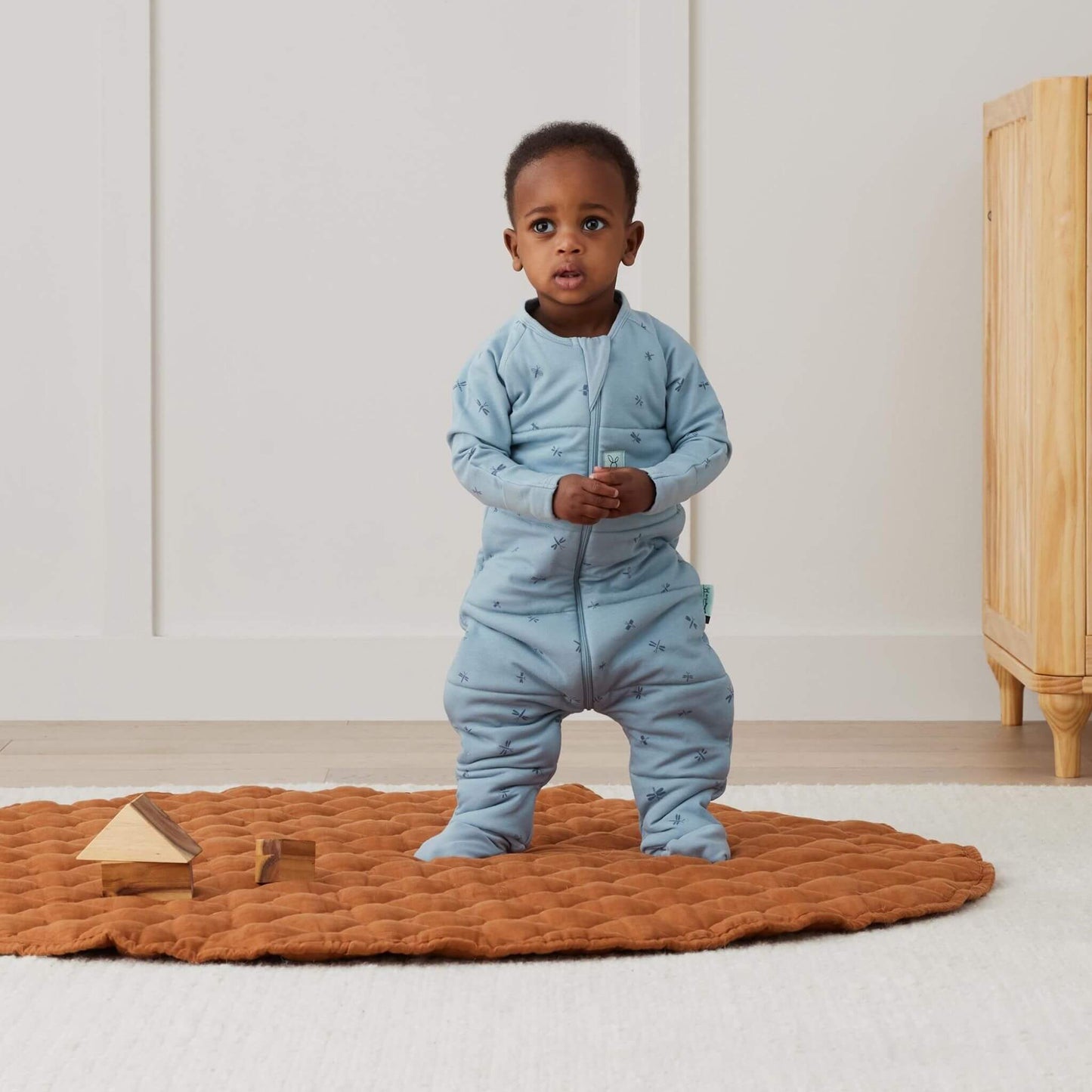 A toddler standing confidently on a quilted rust-coloured mat, dressed in a blue Sleep Onesie in Dragonflies print.
