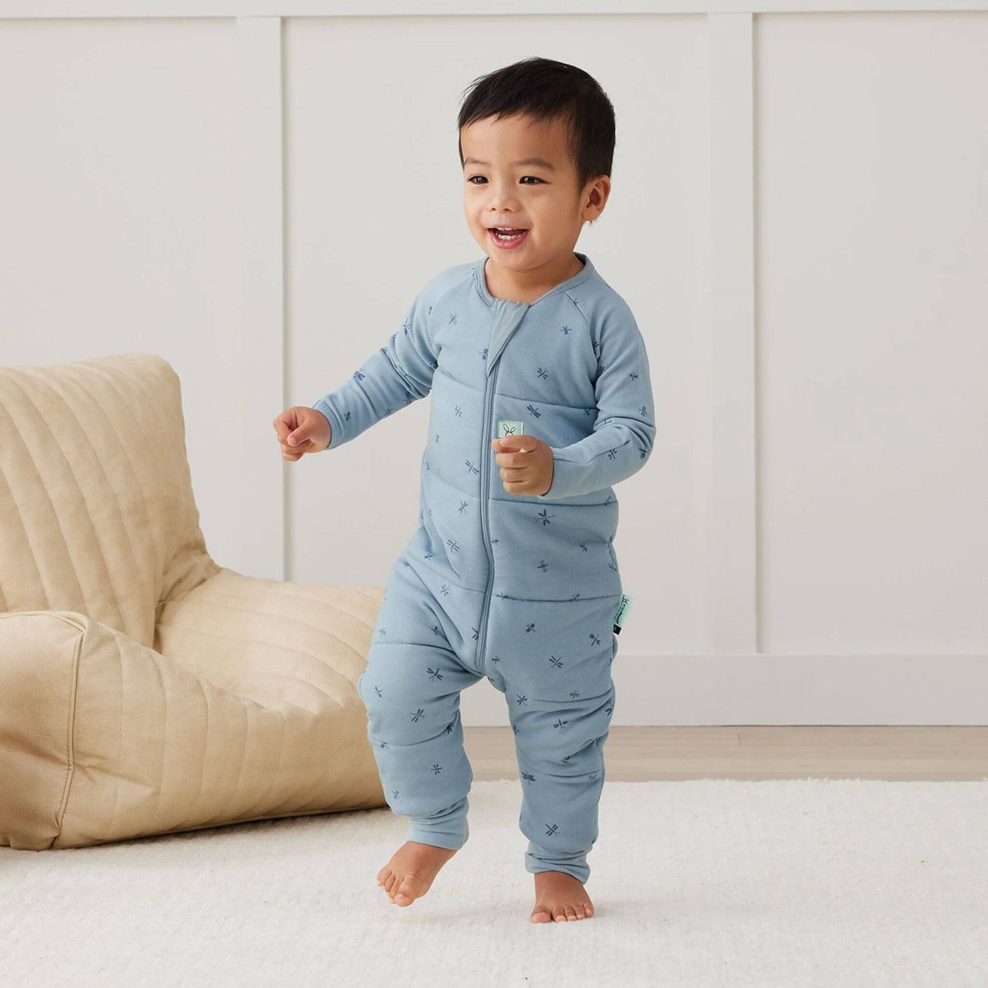 A smiling toddler joyfully walking across a cosy indoor space, dressed in a blue winter weight Sleep Onesie in Dragonflies print. 