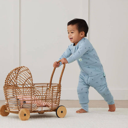 A happy toddler pushes a wicker toy pram while wearing a blue Sleep Onesie in Dragonflies print.