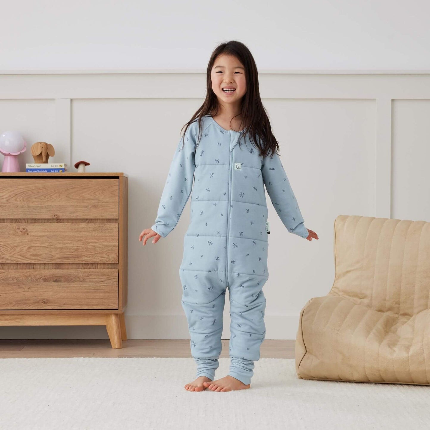 A cheerful child stands wearing a blue Sleep Onesie in Dragonflies print, showcasing its snug, cosy design and flexible fit.