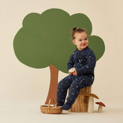 A toddler wearing a Sleep Onesie in Hedgehog print sits comfortably on a wooden stump, surrounded by playful woodland-themed decor, including a tree backdrop and felt mushrooms.