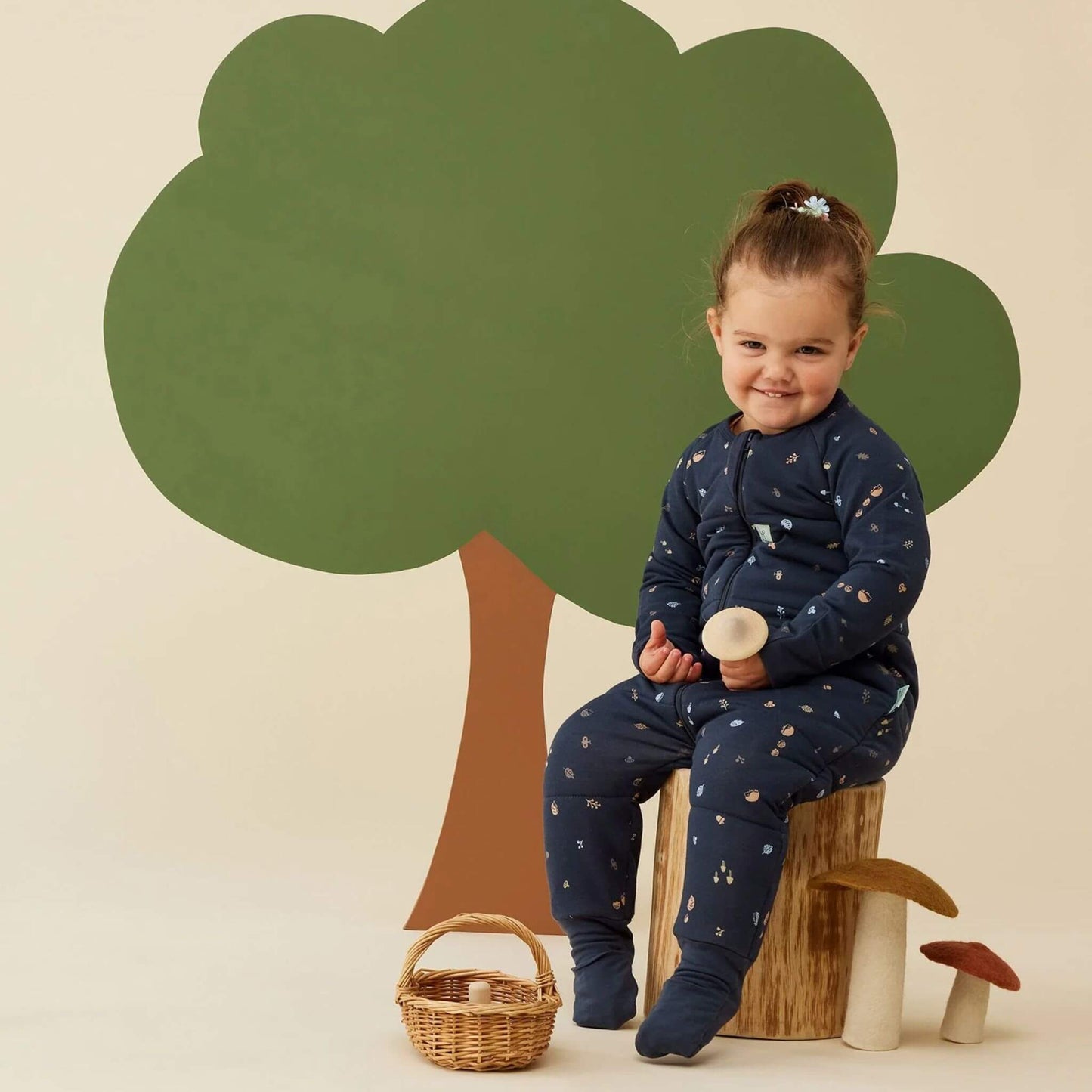A toddler smiles warmly while seated on a wooden stump, wearing a cosy winter weight Sleep Onesie in Hedgehog print. 