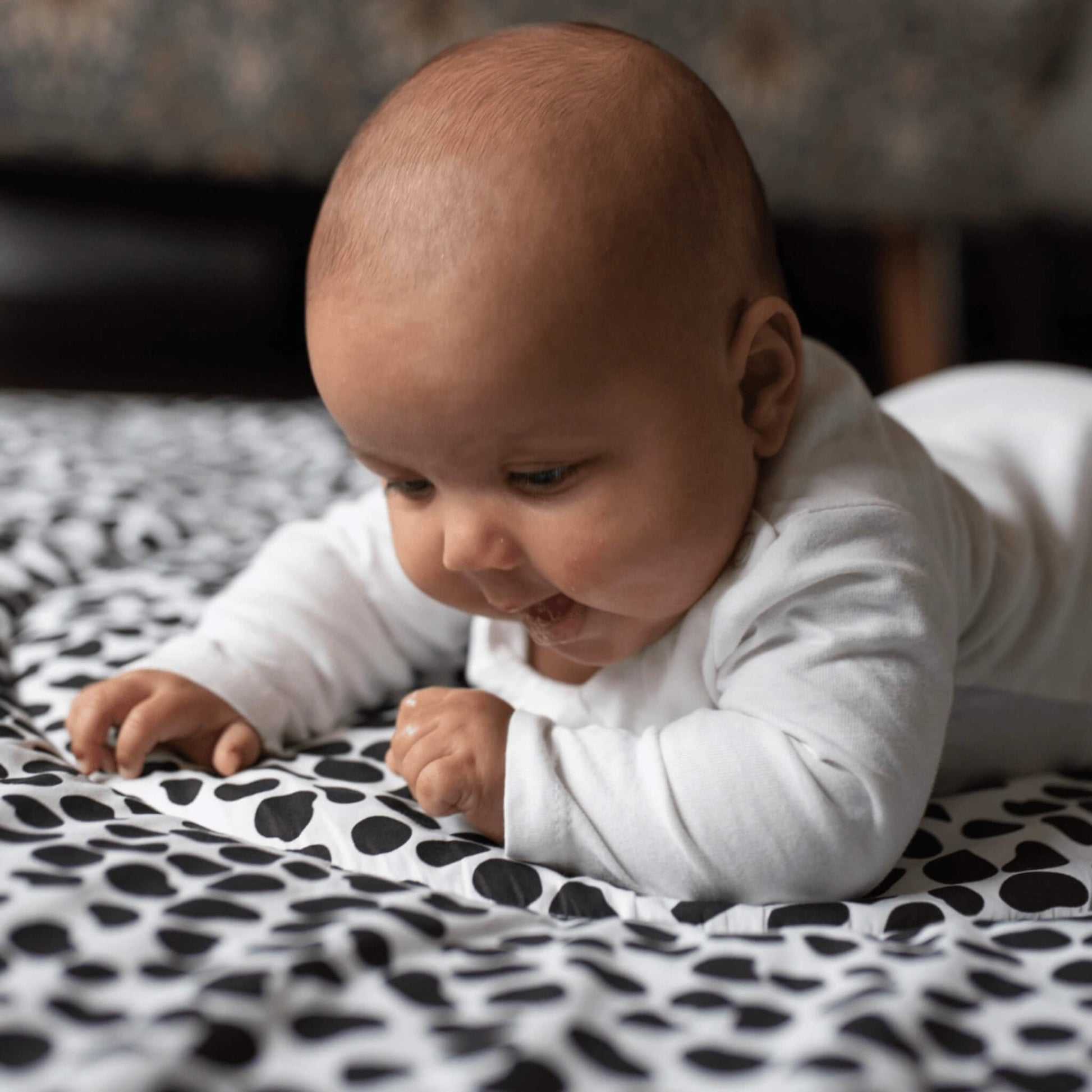 Baby lying on tummy on a padded black and white patterned playmat, focusing closely on the bold high contrast shapes during floor play.