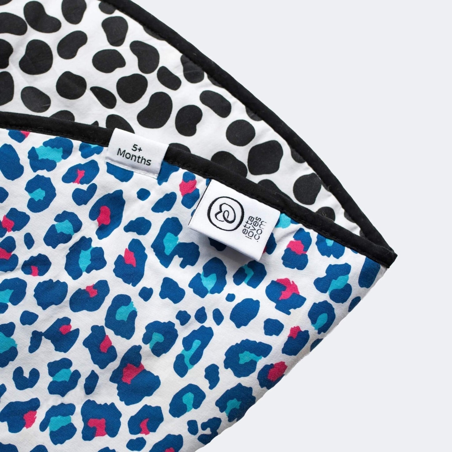 Close up of a reversible playmat corner showing colourful leopard print fabric, stitched edging and woven age label details.