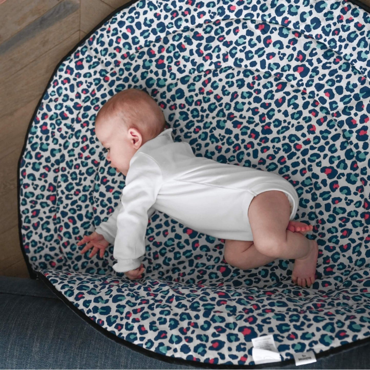 Baby practising tummy time on a soft padded mat with blue and pink leopard print pattern, positioned on a wooden floor.
