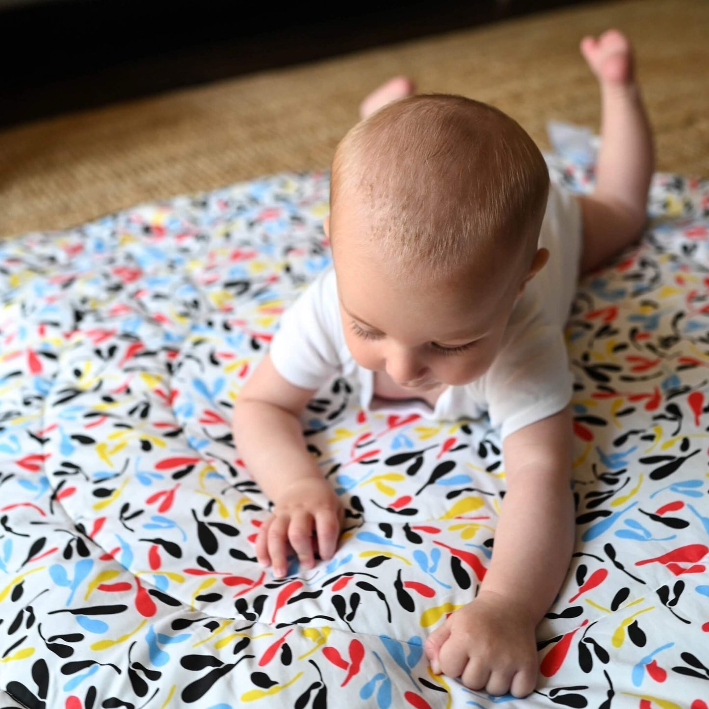 Baby practising tummy time on colourful abstract side of padded sensory mat with red, blue, yellow and black shapes.