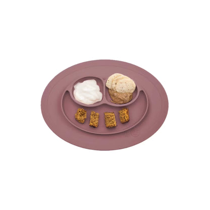 Mauve silicone suction placemat with three divided sections filled with yogurt, banana slices, and small baked bread portions arranged for a toddler meal.