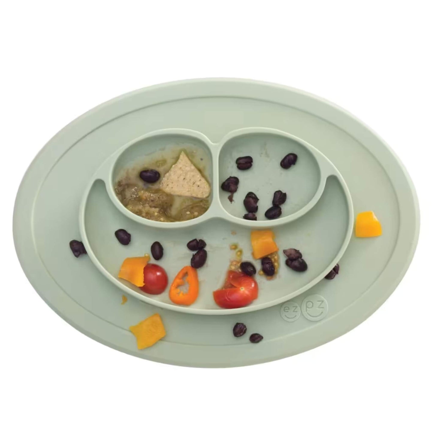 Sage green silicone suction placemat with three divided sections containing leftover food including beans, tomato, pepper pieces, and a tortilla chip.
