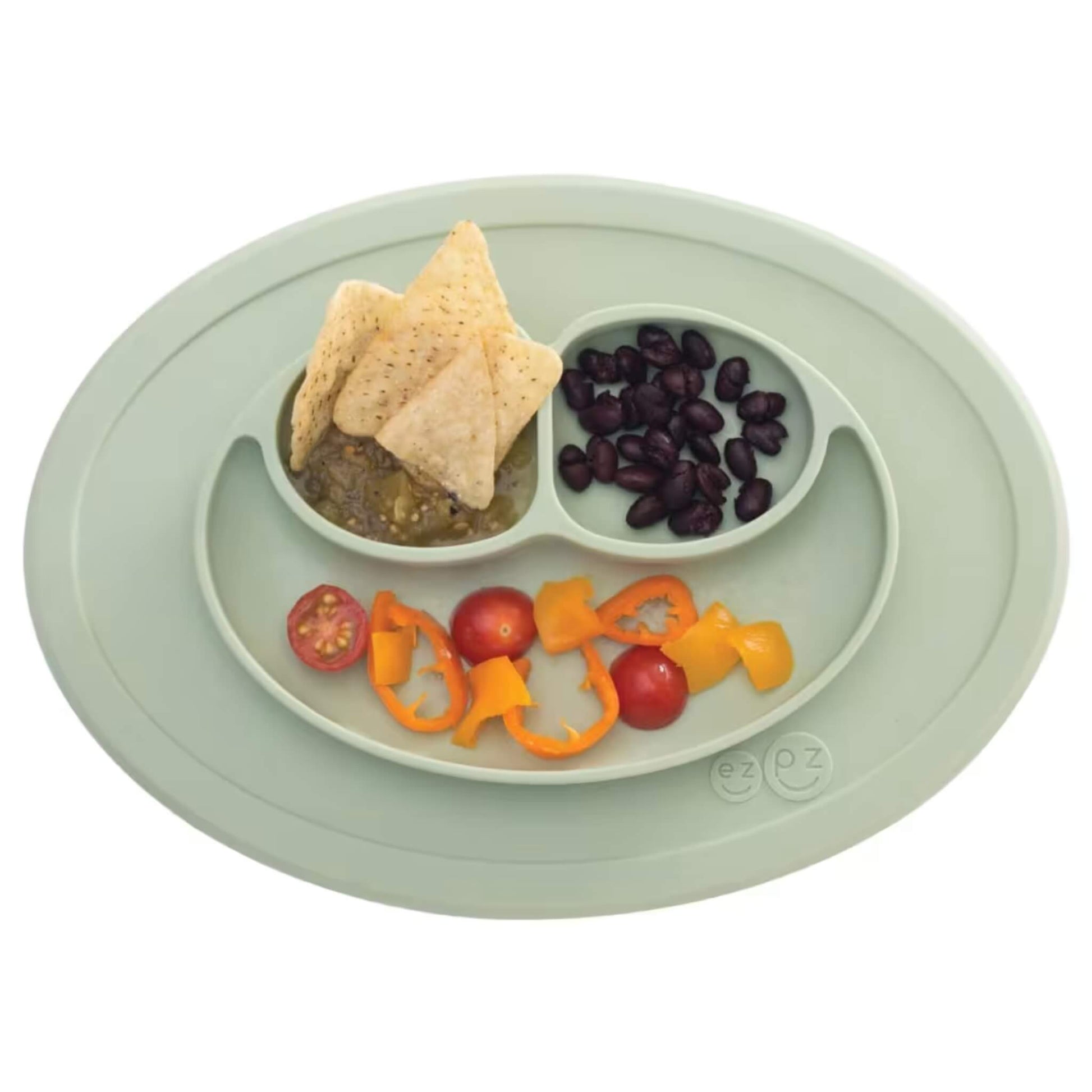 Silicone suction placemat in sage green shown with food portions, including tortilla chips, black beans, cherry tomatoes, and sliced peppers arranged in sections.