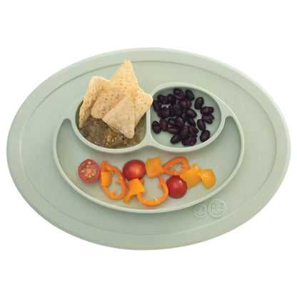 Silicone suction placemat in sage green shown with food portions, including tortilla chips, black beans, cherry tomatoes, and sliced peppers arranged in sections.