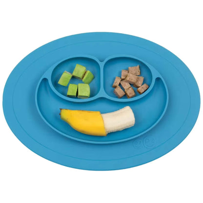 A blue silicone suction baby weaning plate holding avocado cubes, bite-size meat pieces and a half banana arranged in the three smile-shaped sections on white.