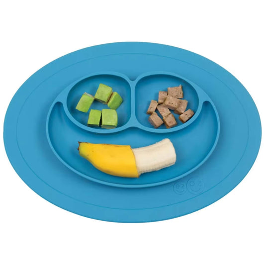 A blue silicone suction baby weaning plate holding avocado cubes, bite-size meat pieces and a half banana arranged in the three smile-shaped sections on white.