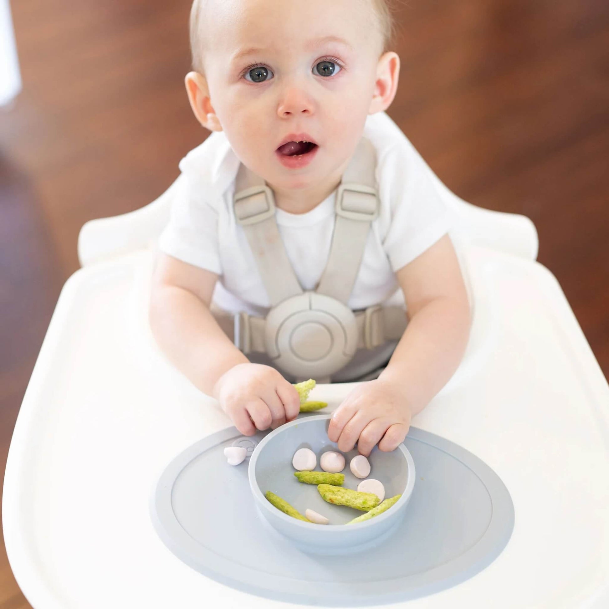 A baby in a white top sitting in a highchair eating snacks from a grey silicone placemat with a small built-in bowl placed on the tray.