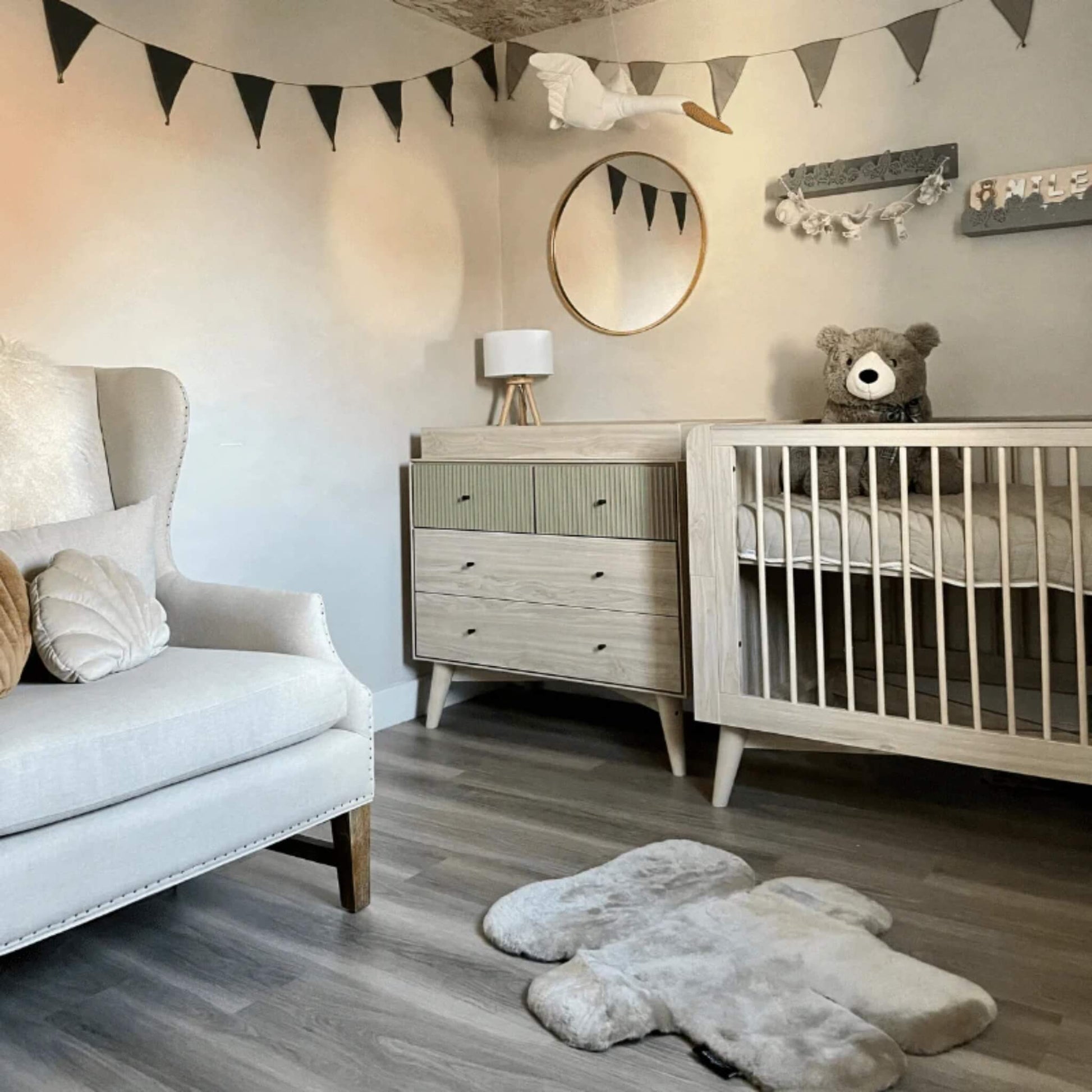 A neutral nursery interior with a bear-shaped sheepskin play rug laid flat on a wooden floor beside a cot, dresser and upholstered chair.