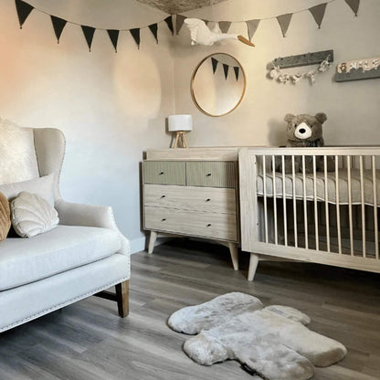 A neutral nursery interior with a bear-shaped sheepskin play rug laid flat on a wooden floor beside a cot, dresser and upholstered chair.