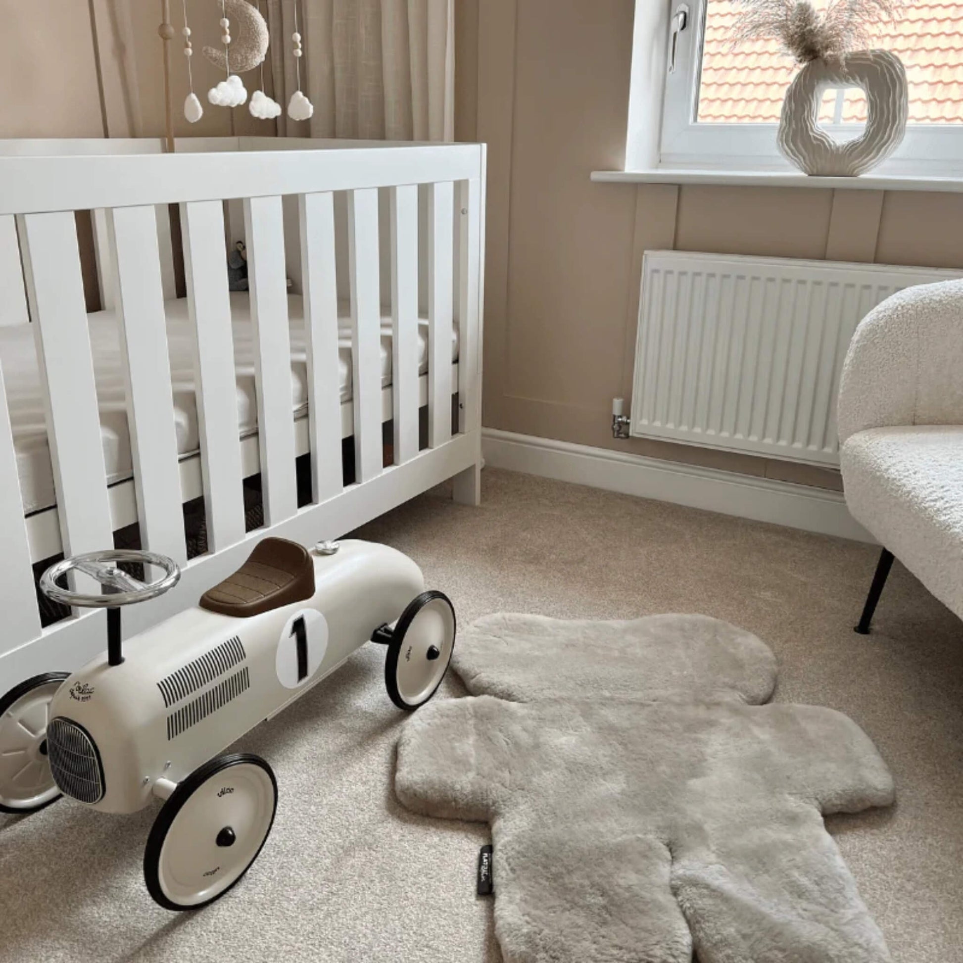 A bear-shaped sheepskin baby play rug placed on carpeted flooring near a cot and a child’s ride-on toy, showing scale and soft texture.